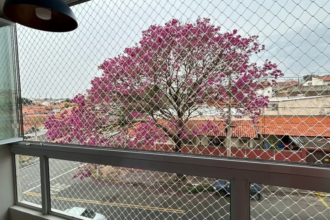 Rede de proteção instalada na janela de um apartamento, proporcionando segurança com vista para a rua e para uma grande árvore com flores rosas.