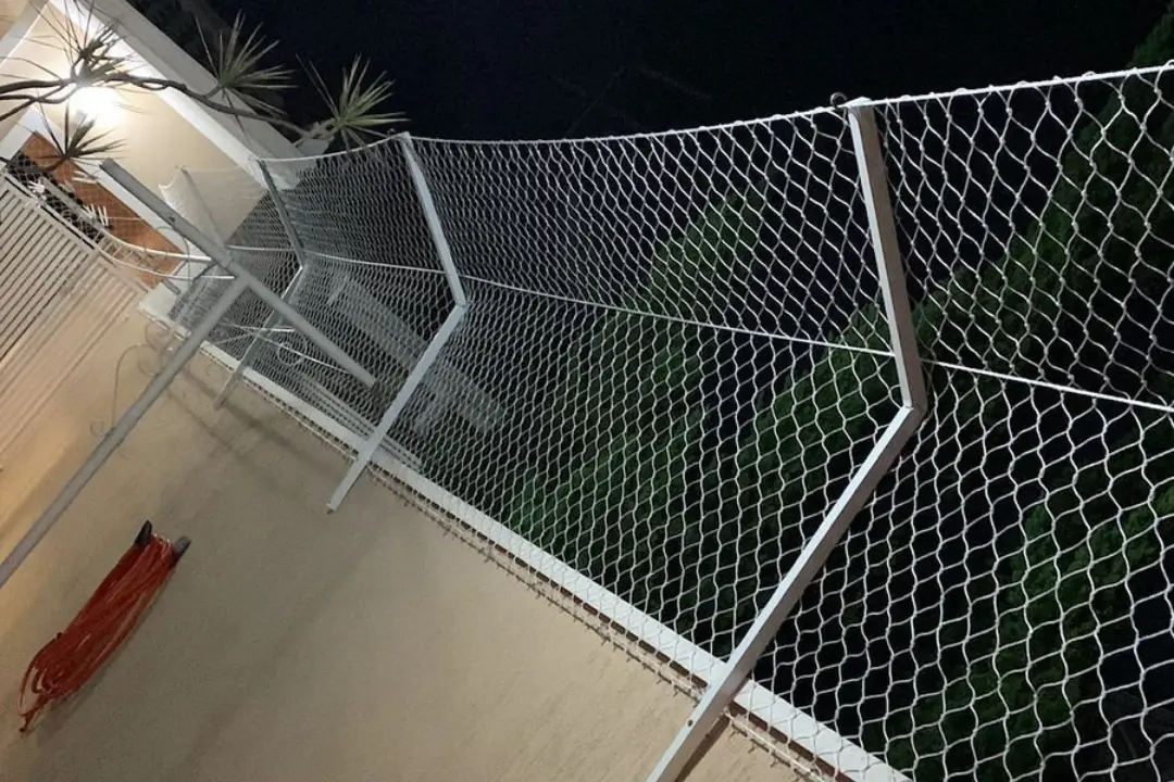 Rede de proteção fixada em uma estrutura metálica branca sobre um muro, fotografada no período noturno, proporcionando segurança extra para a residência.