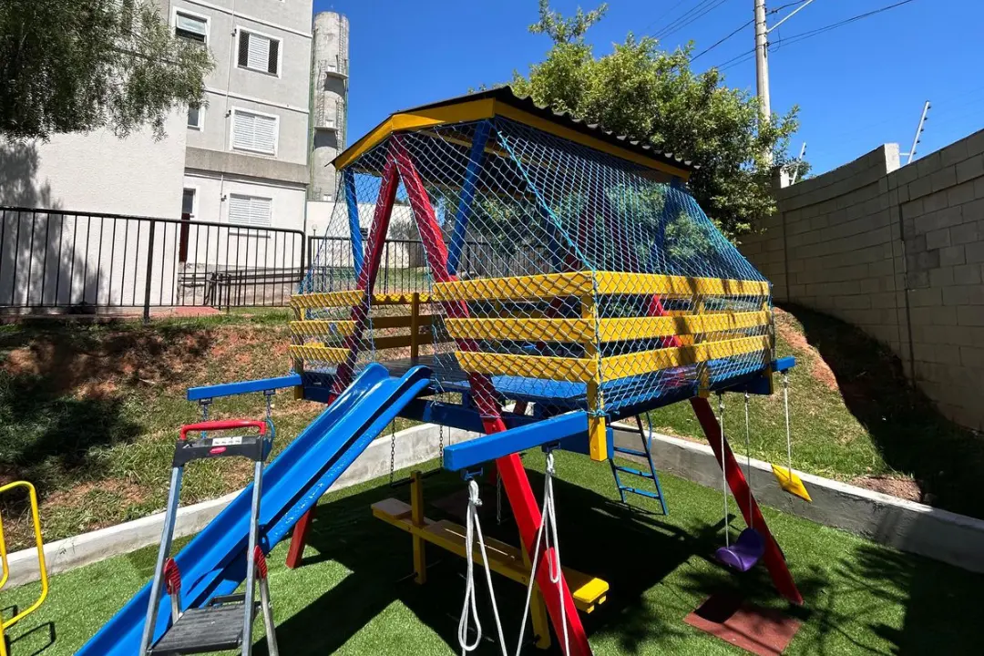 Playground infantil de madeira e metal com escorregador localizado em uma área externa, envolvido por rede de proteção para evitar quedas.