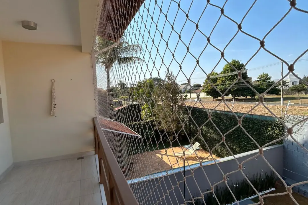 Sacada de uma casa com guarda-corpo de madeira, fechada com rede de proteção tensionada, com vista para uma área externa ensolarada.